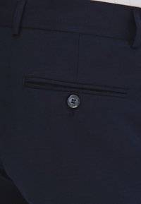Granatowe spodnie o gładkiej teksturze, z tkaną strukturą, boczną kieszenią z zapięciem na guzik oraz subtelnymi detalami w przeszyciu.