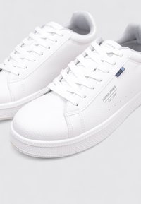 Zapatillas blancas de cuero con superficie texturizada, puntera redonda, cordones blancos planos y detalles del logo en azul. Suela de goma con dibujo en el patrón.