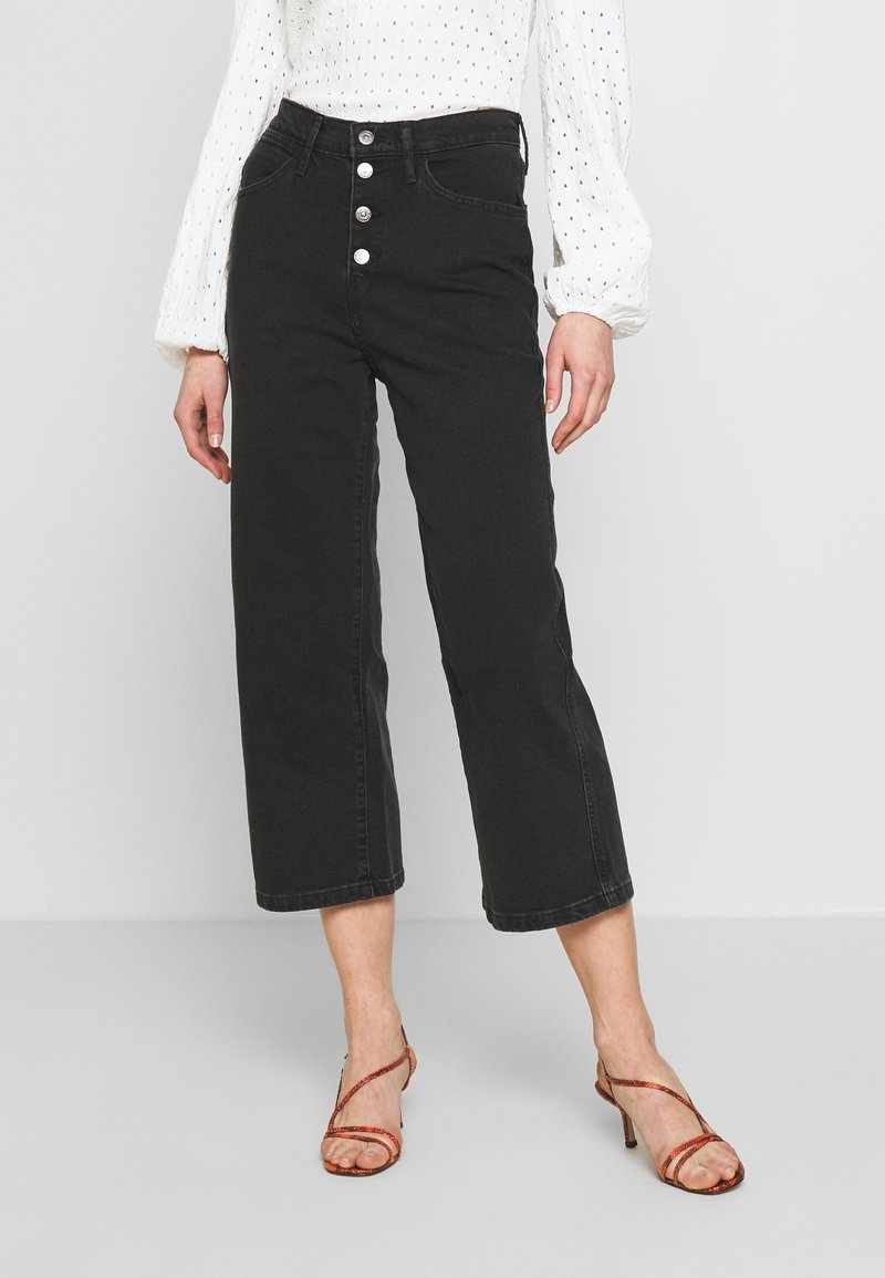 Femme portant un jean noir court taille haute avec des boutons argentés, une blouse blanche à manches longues et des talons orange à brides sur un sol gris.
