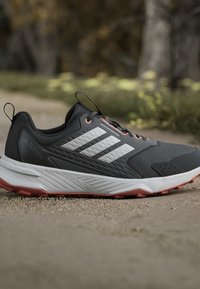 Chaussure de course Adidas Terrex noire et grise avec des bandes blanches et des accents orange sur un sentier extérieur pavé.