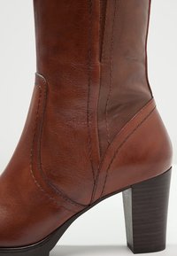 Stivale alla caviglia in pelle marrone con zipper verticale, cuciture curve e tacco largo in legno. Texture liscia con una sottile lucentezza.