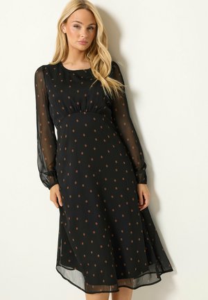 Blonde Frau trägt ein schwarzes Midikleid mit braunen Polka-Dots und transparenten langen Ärmeln, steht vor weißem Hintergrund.