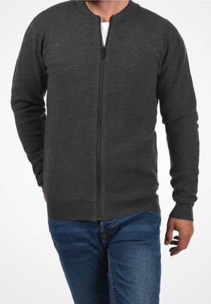 Mężczyzna ubrany w ciemnoszary prążkowany sweter na zamek błyskawiczny na biały shirt i niebieskie dżinsy, pokazany od szyi do bioder na białym tle.