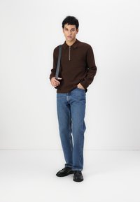 Polo marron à manches longues avec un col zippé, associé à un jean en denim bleu clair. Le modèle se tient debout avec des chaussures noires sur un fond blanc.