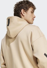 Sweatshirt bege com um tecido texturizado, apresentando um capuz grande e um detalhe de logo preto nas costas. Design suave e casual com ombros caídos.