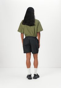 Olive green oversized shirt, black shorts with elastic waistband, white ankle socks, and dark sneakers. Smooth texture, minimal design.