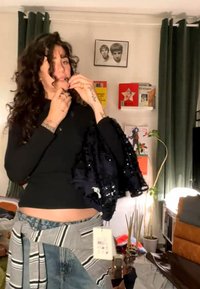 Kvinde med krøllet hår iført sort top og jeans, der holder en sort palietjakke i et rodet rum med bøger, planter og gardiner.