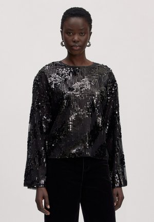 Schwarze Frau trägt ein lockeres schwarzes Langarmoberteil, bedeckt mit großen glänzenden schwarzen und silbernen Pailletten, kombiniert mit schwarzen Hosen.