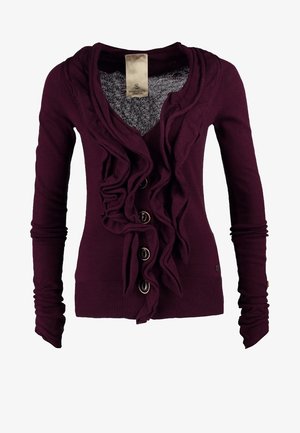 Bordowy sweter rozpinany z falbanami i guzikami, zestawiony z niebieskimi dżinsami i czarną torebką. Modelka nosi czarne szpilki w szpic.