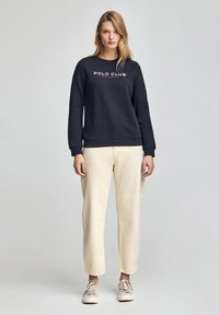 Felpa girocollo navy con testo "POLO CLUB" in bianco e rosso. Abbinata a pantaloni beige chiaro a gamba larga e sneakers beige.
