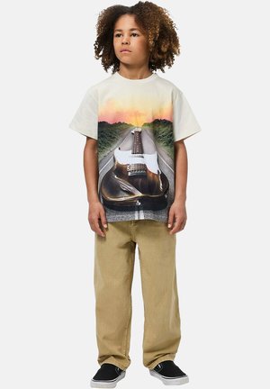 Maglietta grafica con una chitarra su una strada con uno sfondo al tramonto. Colore crema chiaro, maniche corte, vestibilità comoda. Abbinata a pantaloni beige.