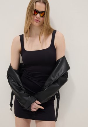 Donna che indossa un abito nero aderente, giacca di pelle appoggiata sulle spalle e occhiali da sole con lenti arancioni, in piedi davanti a un muro chiaro e semplice.