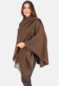 Frau in einem braunen, fransigen Wollponcho über schwarzen Leggings, die vor einem einfarbigen, hellen Hintergrund steht und leicht nach rechts schaut.