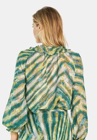 Blusa a maniche lunghe con colletto a volant e motivo a righe verticali verdi, gialle e blu su materiale leggero.