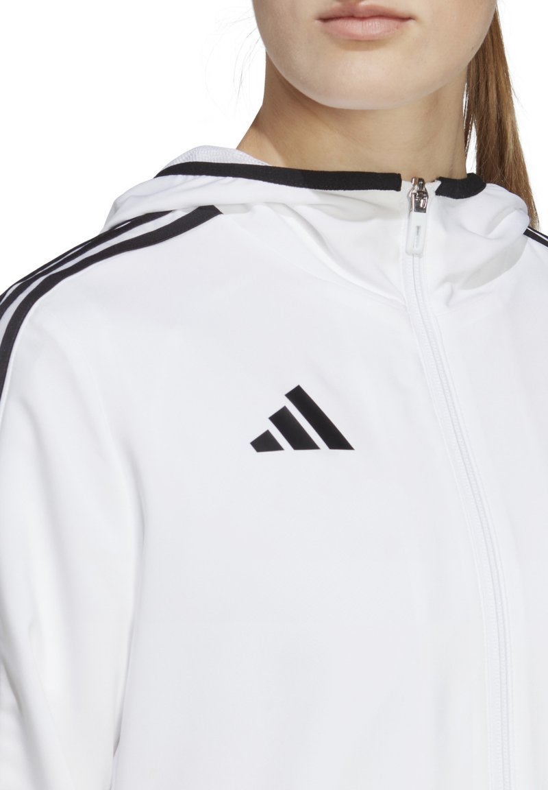 Giacca a zip bianca con dettagli neri e design a tre strisce sulle maniche. Presenta un logo Adidas sul lato sinistro del petto. Tessuto liscio e testurizzato.