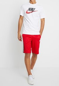 White Nike t-shirt with black and red logo, paired with red athletic shorts featuring black side stripes and white sneakers.