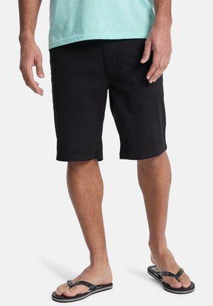 Mann trägt schwarze Shorts, hellblaues Shirt und schwarze Flip-Flops, steht vor einem einfarbigen Hintergrund.