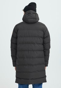 Svart polstret parka med hette, med et kviltet tekstur og en midtlengdes design. Bakview viser et glatt stoff og minimale detaljer.