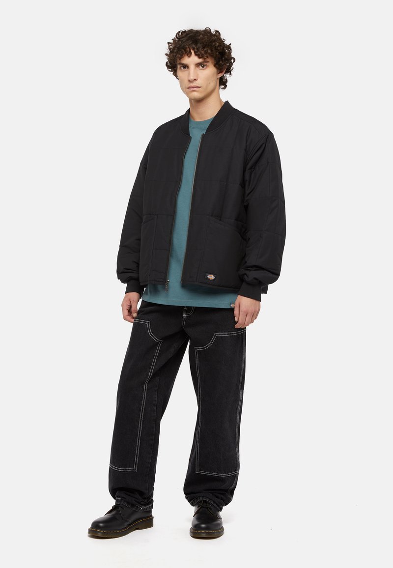 Veste bomber noire matelassée avec des poignets côtelés, assortie à un t-shirt bleu sarcelle à col rond et un jean noir ample avec des coutures blanches. Des chaussures noires complètent le look.