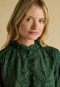 Dunkelgrüne Bluse mit Rüschenausschnitt, die über florale Stickereien und eine Knopfleiste verfügt, aus weichem Stoff mit strukturierter Oberfläche.