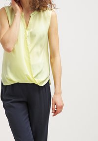 Sleeveless yellow blouse with a V-neck and draped design, made of lightweight, smooth fabric. Paired with dark navy trousers.