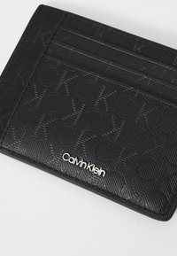 Porte-cartes en cuir noir avec un motif géométrique en relief, doté de plusieurs emplacements pour cartes et d'un détail en logo argenté. Texture lisse, forme compacte.