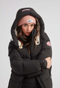 Chaqueta acolchada negra con una gran capucha, forro color crema y un parche de logo de goma texturizada. Presenta puños elásticos y bolsillos con cremallera.