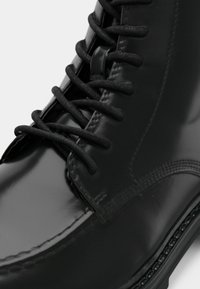 Črni škornji iz lakiranega usnja z zavezovanjem na vezalke, gladko teksturo, okroglo konico in šivi ob podplatu.