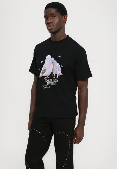 Svart bomull t-shirt med en grafik av två duvor i pastellfärger och hjärtan, texten lyder "Från Paris Med ♥" under.