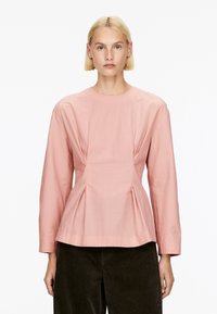 Rosa Bluse mit langen Ärmeln und rundem Ausschnitt, versehen mit einem gefältelten Design an der Taille und glattem, leichtem Stoff. Kombiniert mit dunklen Cordhosen.