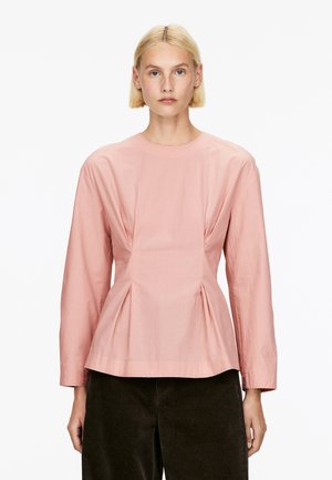 Rosa Bluse mit langen Ärmeln und rundem Ausschnitt, versehen mit einem gefältelten Design an der Taille und glattem, leichtem Stoff. Kombiniert mit dunklen Cordhosen.