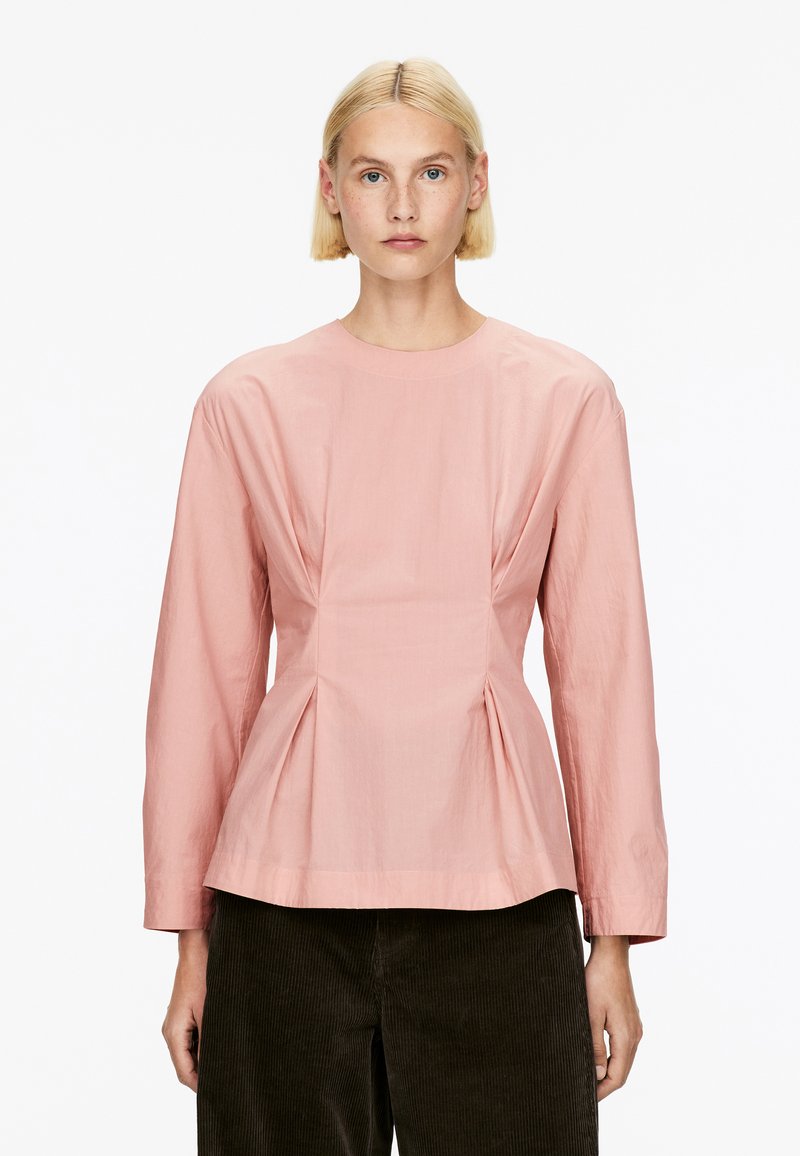 Rosa Bluse mit langen Ärmeln und rundem Ausschnitt, versehen mit einem gefältelten Design an der Taille und glattem, leichtem Stoff. Kombiniert mit dunklen Cordhosen.