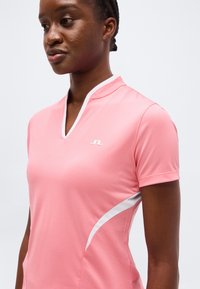 Γυναίκα που φοράει εφαρμοστό ροζ μπλουζάκι με κοντό μανίκι και λευκή επένδυση με μικρό λογότυπο, κοιτάζοντας ελαφρώς αριστερά σε απλό φόντο.