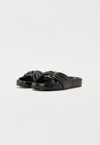 Sandales glissantes en cuir noir avec un design de lanière torsadée, semelle plate et texture lisse. Pas de quincaillerie ou de motifs visibles.