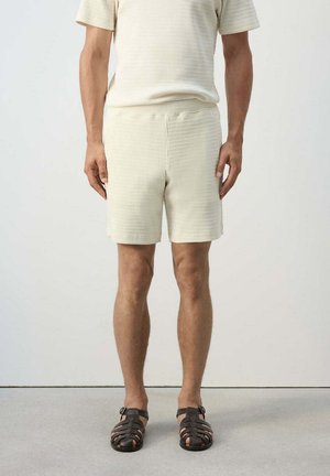 Hombre con pantalones cortos texturizados de color crema y top de manga corta a juego con sandalias de cuero tejidas marrón oscuro, de pie sobre un fondo liso.