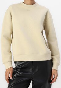 Felpa in cotone beige con collo rotondo, polsini e orlo a coste, caratterizzata da un logo sottile sul petto. Indossata con pantaloni in pelle nera.