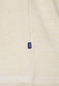 Helles beiges Strickgewebe mit strukturierter Oberfläche, versehen mit einem kleinen dunkelblauen Etikett, das an der Seite angenäht ist. Einfaches, sauberes Design ohne Muster.