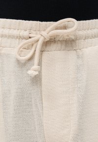 Κοντινή λήψη παντελονιού από ύφασμα σε εκρού χρώμα με υφή, με ελαστική μέση και δεμένο κορδόνι μπροστά σε φιόγκο.