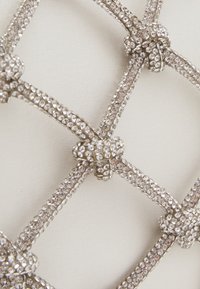 Close-up van een zilveren strassteentjes rasterpatroon met geknoopte kruisingen op een gladde beige stoffen achtergrond.