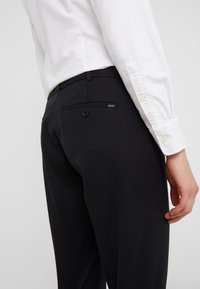 Svarta dressbyxor med en slät textur, med en bakficka med knappstängning och en liten "Polo"-etikett. Vit skjorta delvis synlig.