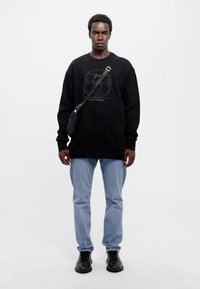 Sudadera negra con un diseño gráfico, combinada con vaqueros azul claro y zapatos negros. Los accesorios incluyen un pequeño bolso cruzado negro.