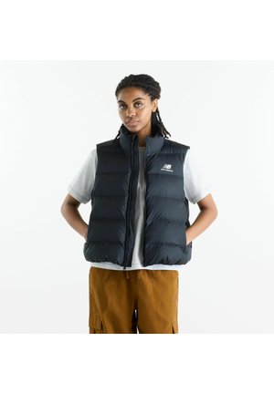 Junge Person mit geflochtenem Haar, die eine schwarze New Balance Steppweste über einem grauen Shirt und brauner Hose trägt, steht mit den Händen in den Taschen.