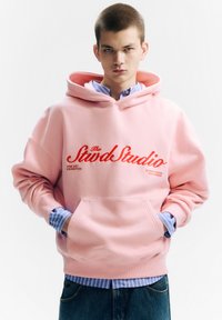 Rózsaszín kapucnis pulóver első zsebével, piros hímzett "The Stwd Studio" logóval, kék-fehér csíkos ing fölött viselve, laza szabásban.