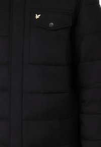 Schwarze Daunenjacke mit gesteppten Abschnitten, einer Klappentasche mit Druckknopfverschluss und einem Emblem auf der Brust. Glatte Textur, leichter Stoff.