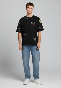 Black oversized T-shirt with graphic designs in red, white, and green, featuring hearts, a butterfly, and text accents. Paired with light blue jeans.
