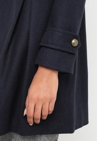 Cappotto in lana blu navy con una texture morbida, dotato di un dettaglio con polsino abbottonato e una vestibilità su misura, mostrato parzialmente con una mano a fuoco.