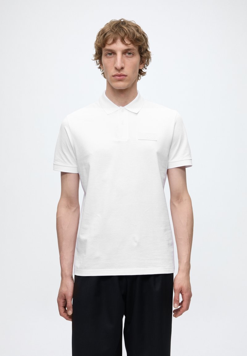 Jeune homme aux cheveux bouclés portant un polo blanc uni à manches courtes et un pantalon noir, debout devant un fond blanc.