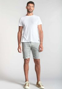 Witte katoenen t-shirt, lichtgrijze cargoshorts met opgerolde zomen, en beige sneakers. Model staat tegen een neutrale achtergrond.