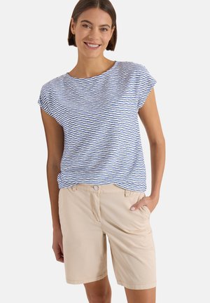 Femme souriante portant un haut blanc et bleu à rayures ondulées à manches courtes et un short beige longueur genoux, main dans la poche, debout devant un fond blanc.