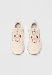 Une paire de chaussures de sport beiges avec des accents bordeaux, des semelles marbrées et des dessus en maille respirante visibles sur un fond blanc.
